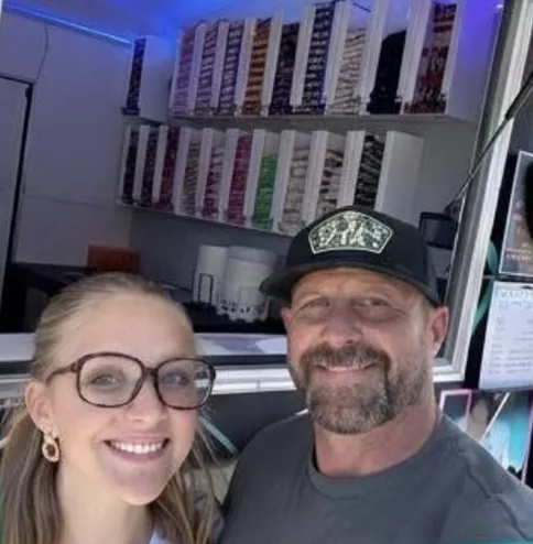 Two of the Noodles Now crew smiling in front of the ramen wall at the service window